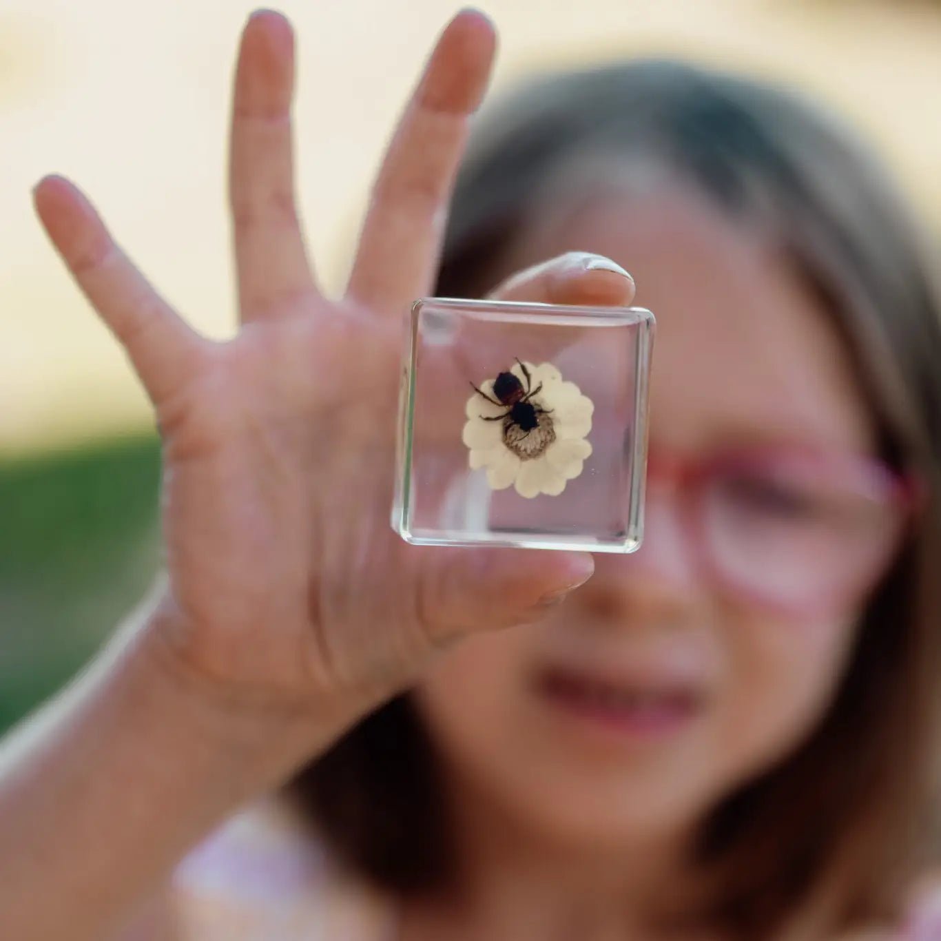 Girl holding a square clear object with a preserved insect and flower, blurred background