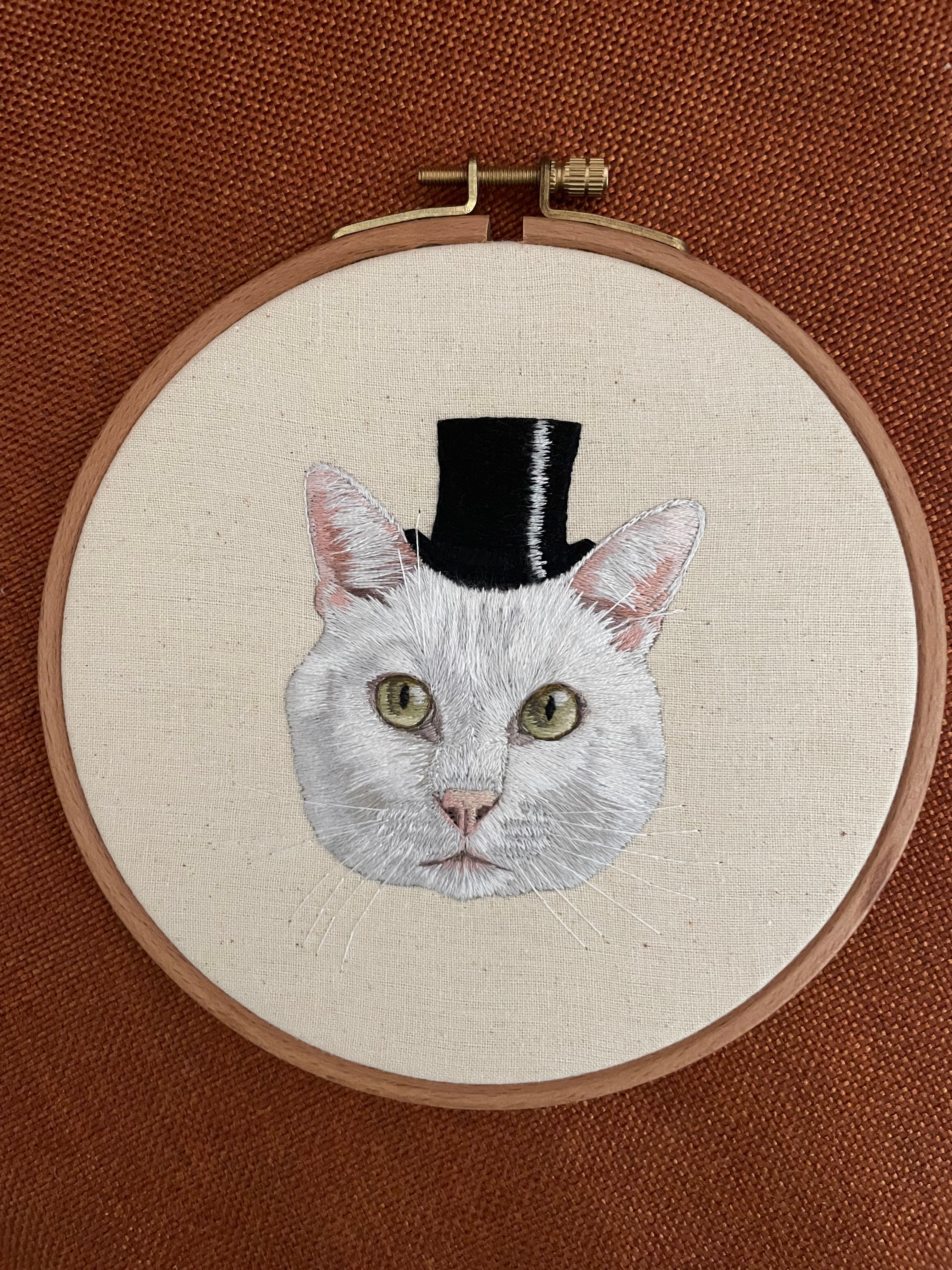 Embroidery of a cat wearing a top hat in an embroidery hoop on a brown background