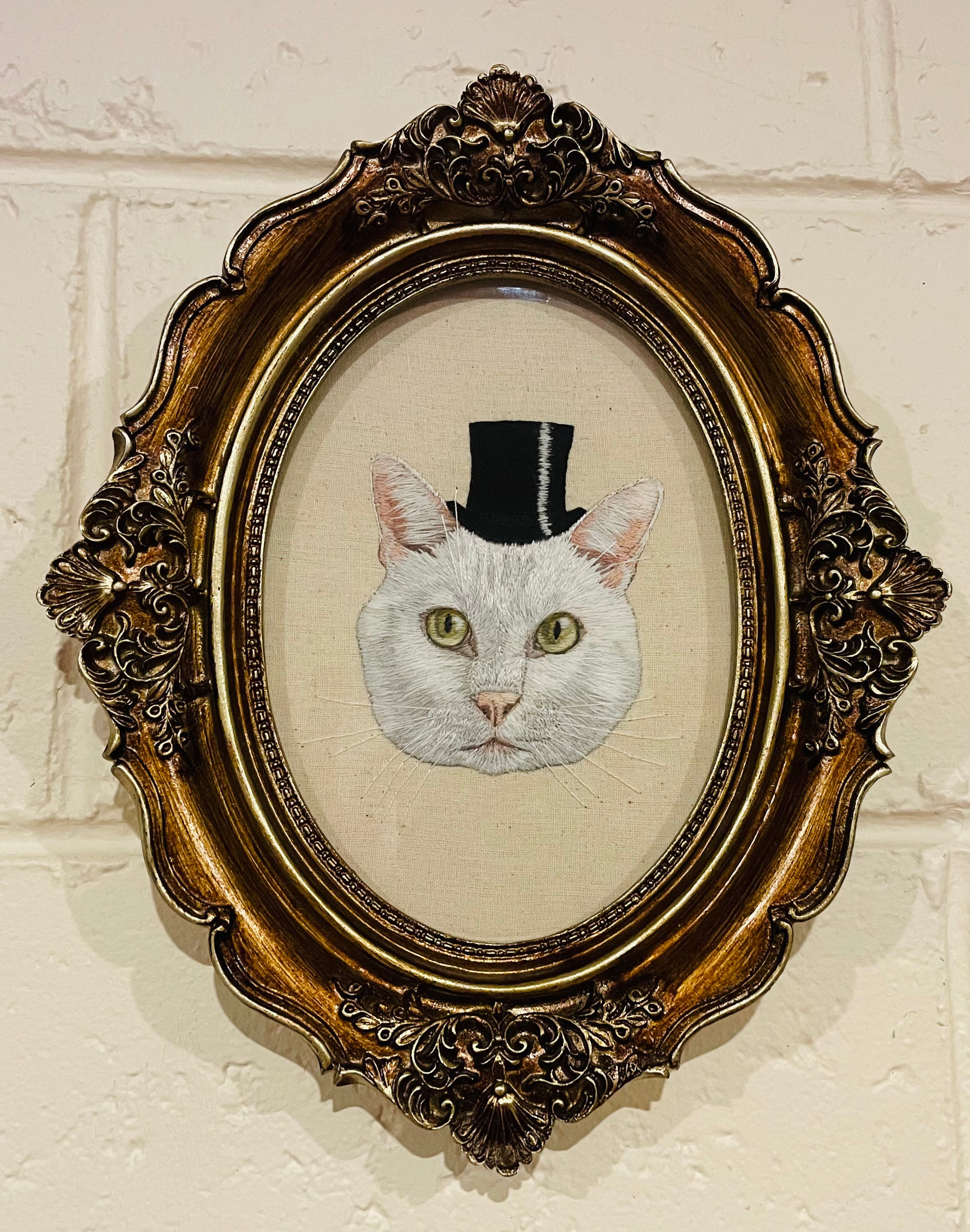 Embroidered picture of a cat wearing a top hat in an ornate gold frame.